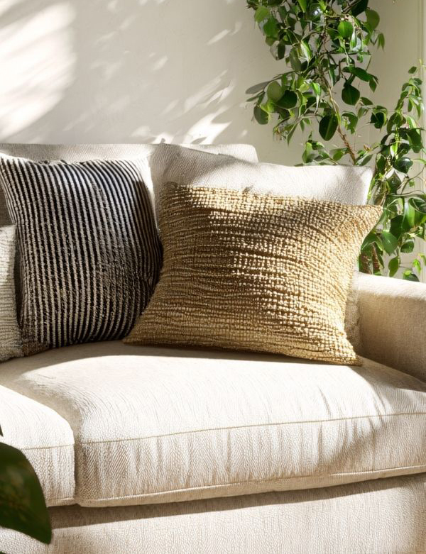 Cream coloured couch with lots of pillows and a plant behind with good light filtering through the window