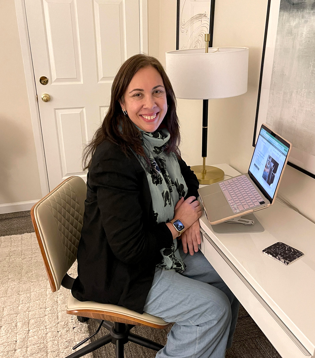 Woman with dark hair sitting at desk with laptop, smiling at the camera
